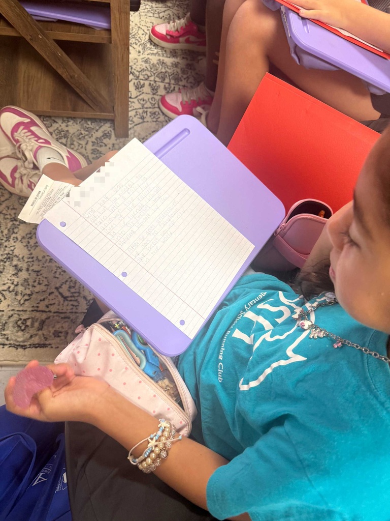 A child reading from a book, seated on the floor. A girl in front is wearing a necklace and bracelets.