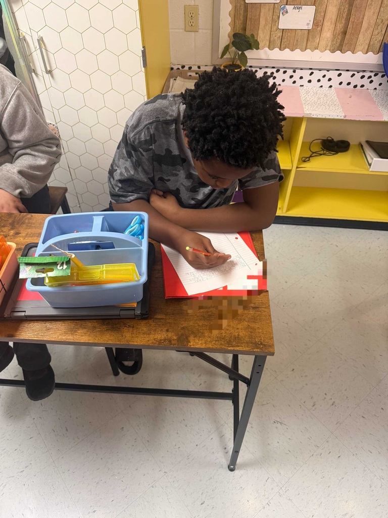 A student wearing glasses leans over a desk, writing on a piece of paper. The desk holds a pencil box.
