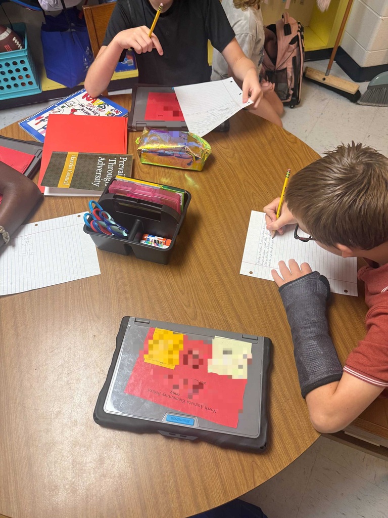Kids seated around a desk, one wearing a cast, both using pencils and paper. Books, colored pencils, and other school supplies are on the desk.