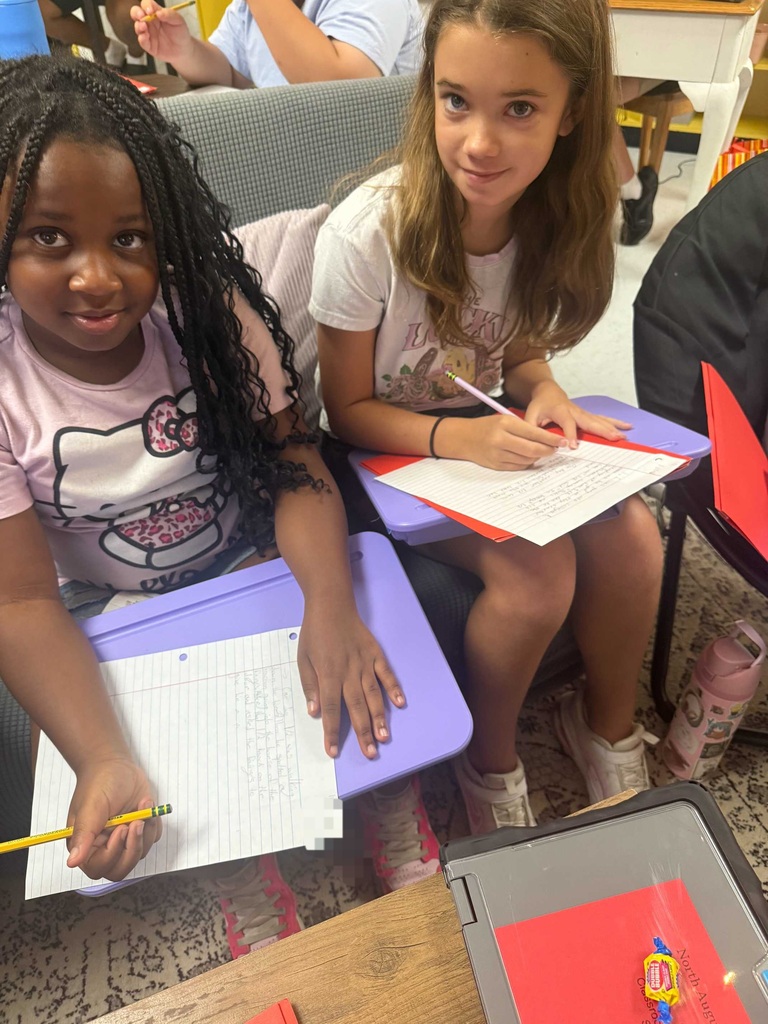 Two young girls seated at desks in a classroom, each holding a pencil and writing on paper.