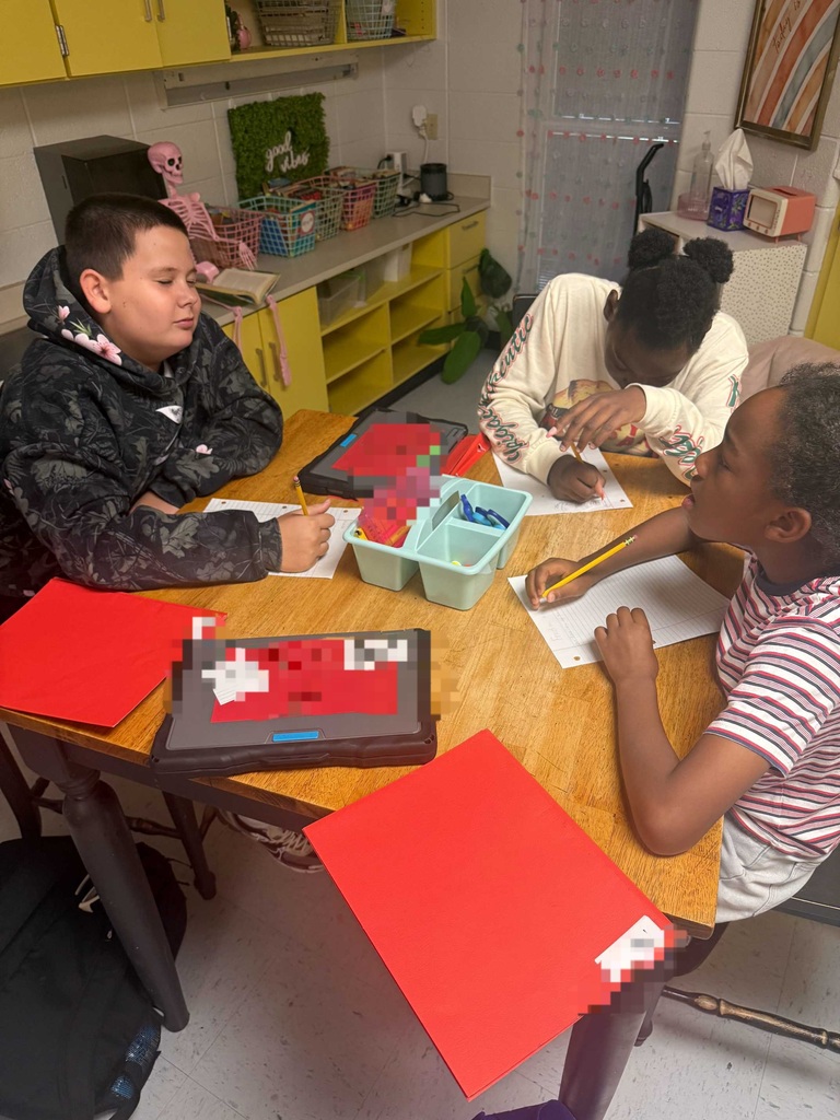 Three children seated around a table, engaged in an activity. One uses a pencil on paper, another holds a tablet, and the third looks at the tablet.