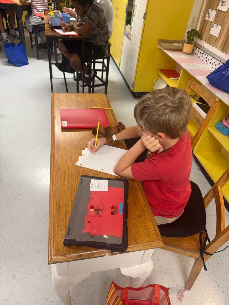 A child in a red shirt sits at a desk, writing on paper with a pencil. Other students sit at tables in a classroom with yellow walls.
