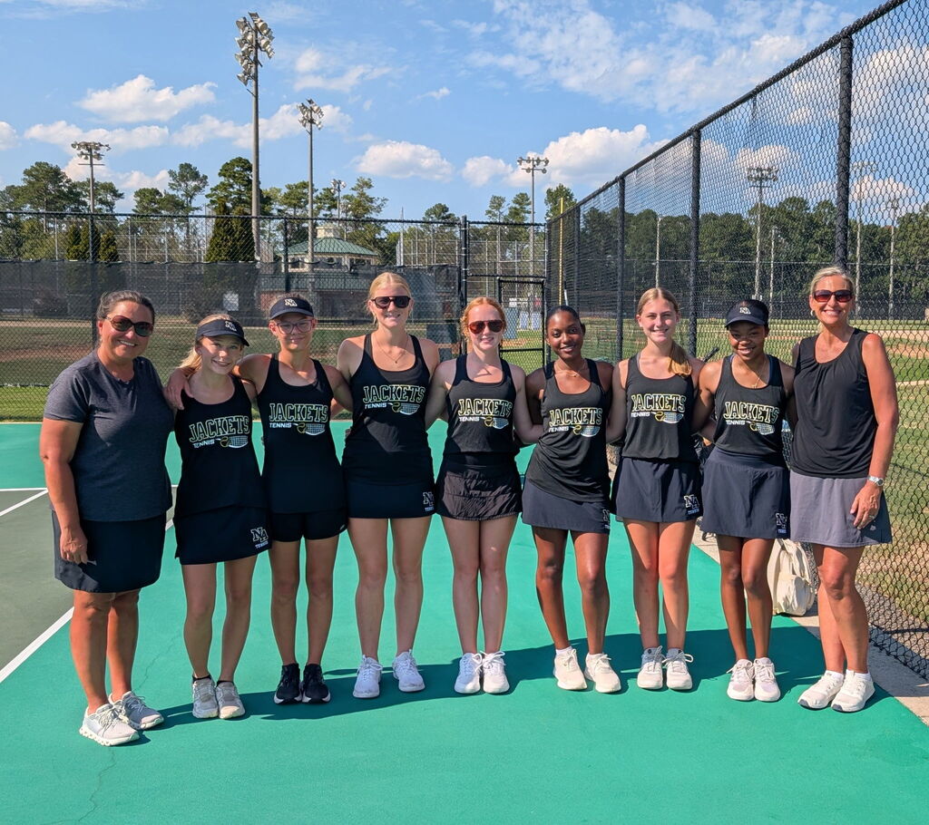 A group of women in matching uniforms stands on a tennis court. Some wear hats. Behind them is a fence, trees, and a clear sky.