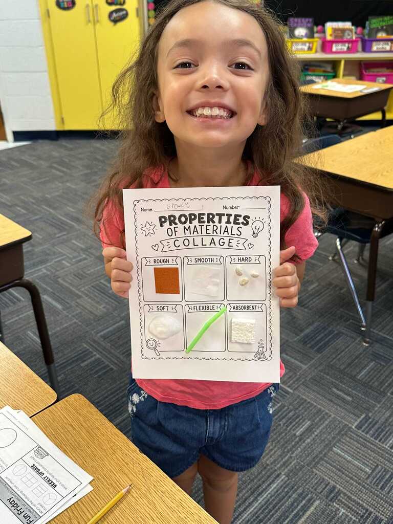 A young girl in a classroom holds a science worksheet titled "Properties of Materials." She smiles with her teeth showing.
