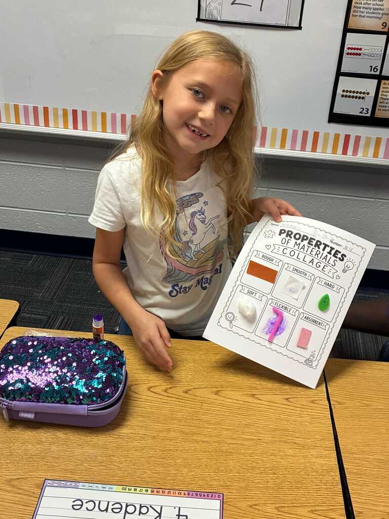 A young girl shows a science poster about material properties. Behind her is a classroom wall with a colorful strip.