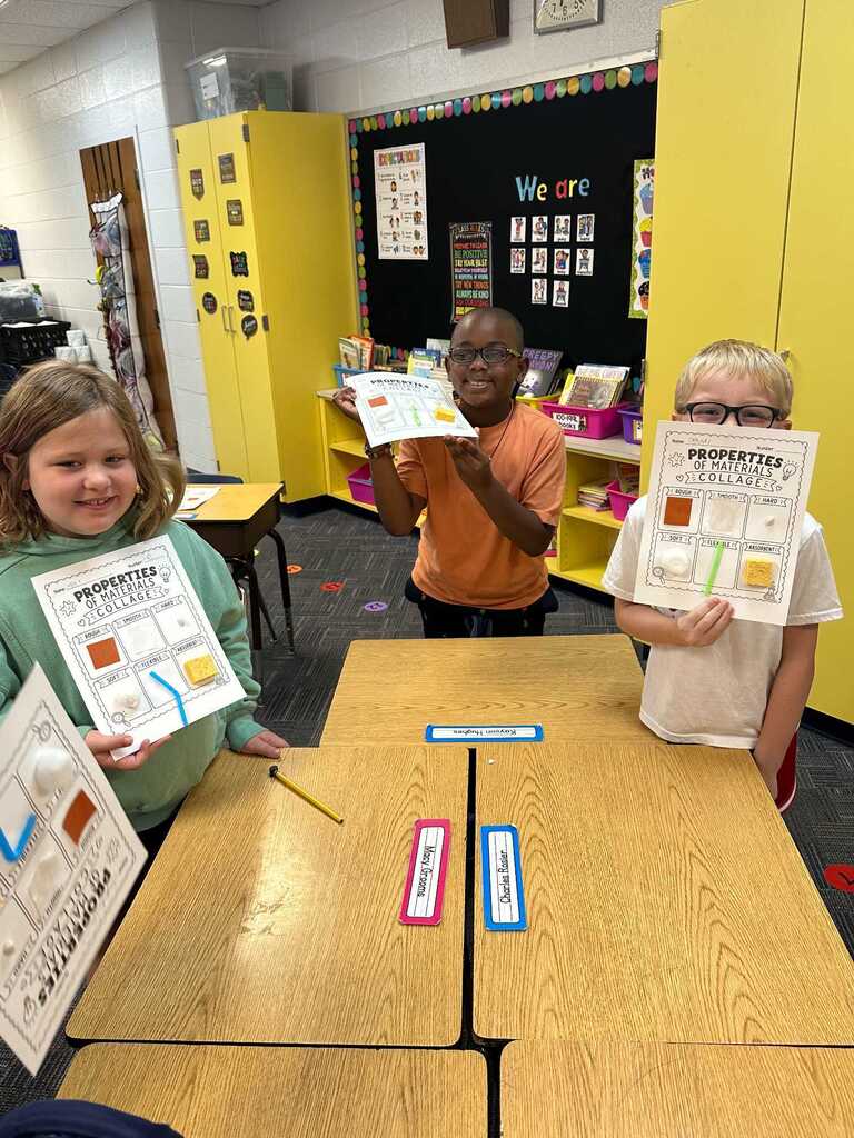 Three children hold up papers with text in a classroom. A table has papers with headings. A yellow wall with a bulletin board is behind them.