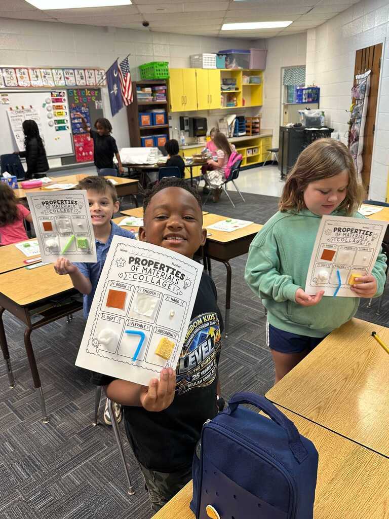 Classroom with desks, shelves, and a flag. Two kids hold up worksheets. One holds a backpack.