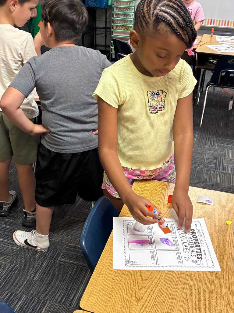 A young girl in a yellow shirt works on an activity at a desk with other children in a classroom.