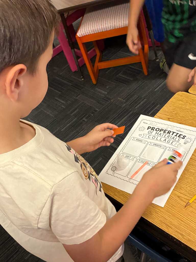 A young boy wearing a white shirt holds a worksheet and orange object in a classroom.