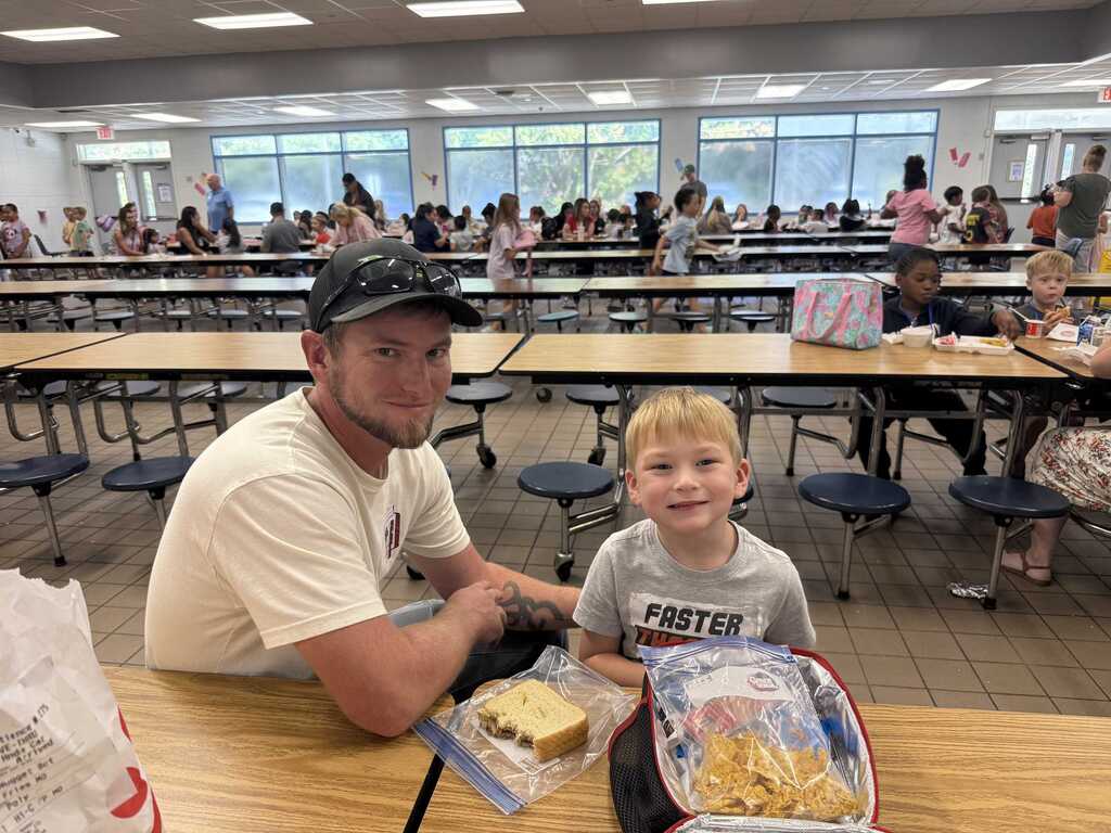 Man and child seated at a table in a school cafeteria, with many other students seated at tables in the background.