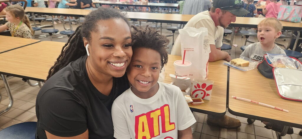 A woman and a boy smile at the camera; they sit together at a cafeteria table.