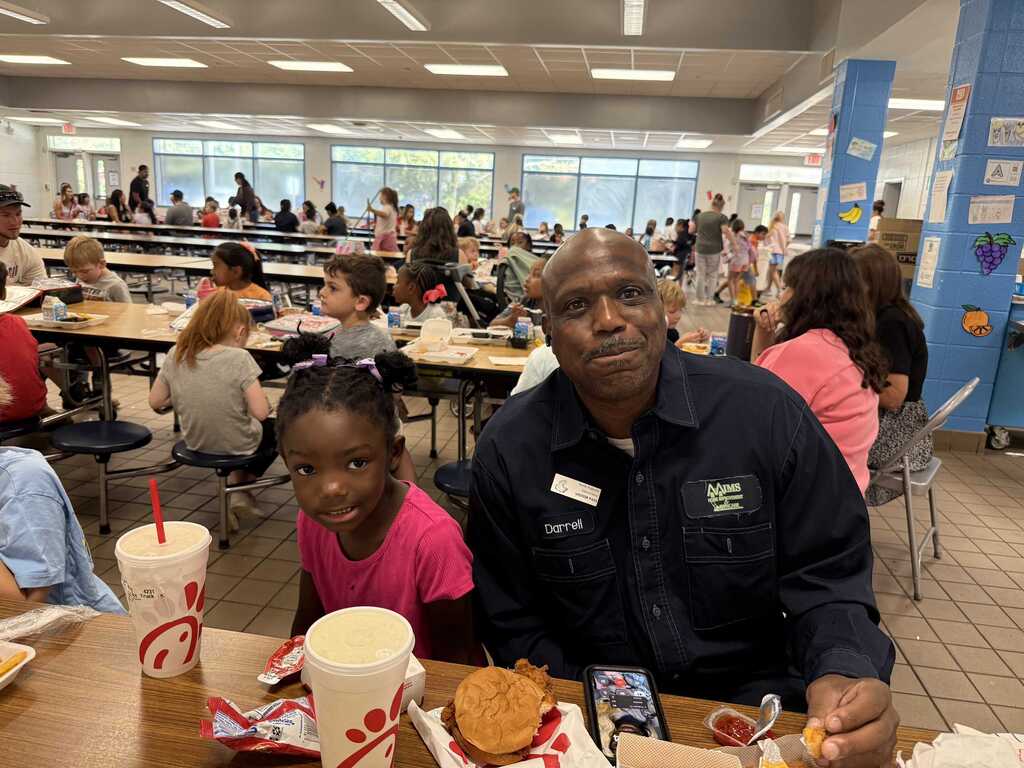 Man and a young girl seated at a table in a cafeteria, eating. Behind them are people seated at tables.