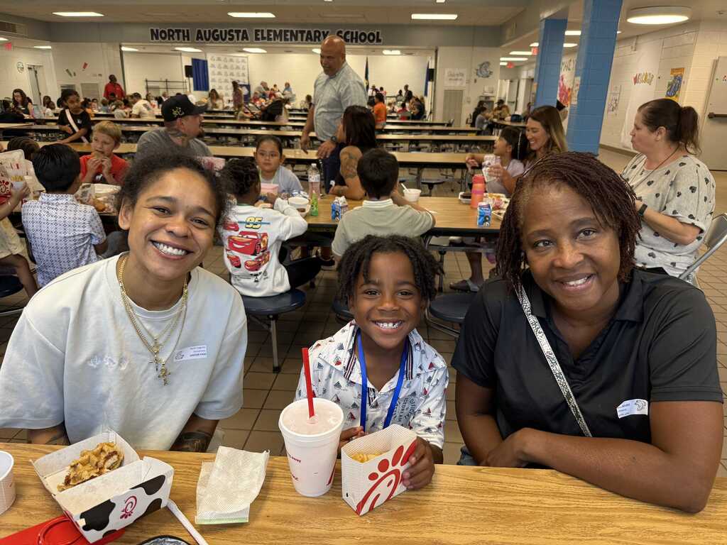 Three people sit at a table, smiling. A girl in the center holds food and drinks. Behind them, a cafeteria with tables and chairs is filled with people eating.