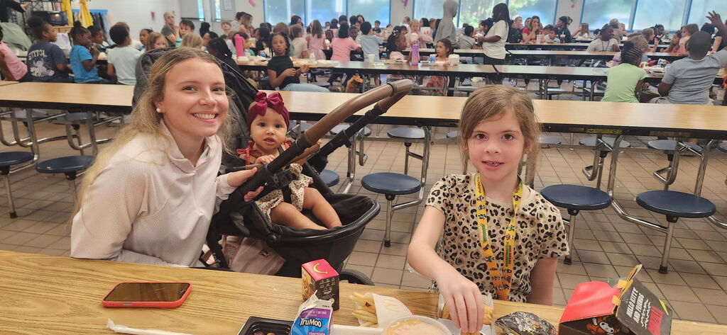 A woman with a baby in a stroller and a young girl with a leopard print shirt sit at a table in a cafeteria.