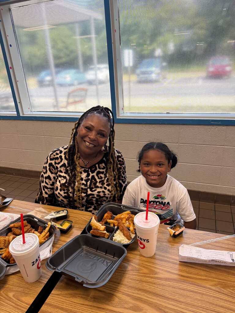 Two individuals seated at a restaurant table, with a tray of food, straws, and drinks. Window in the background shows a parking lot with cars.