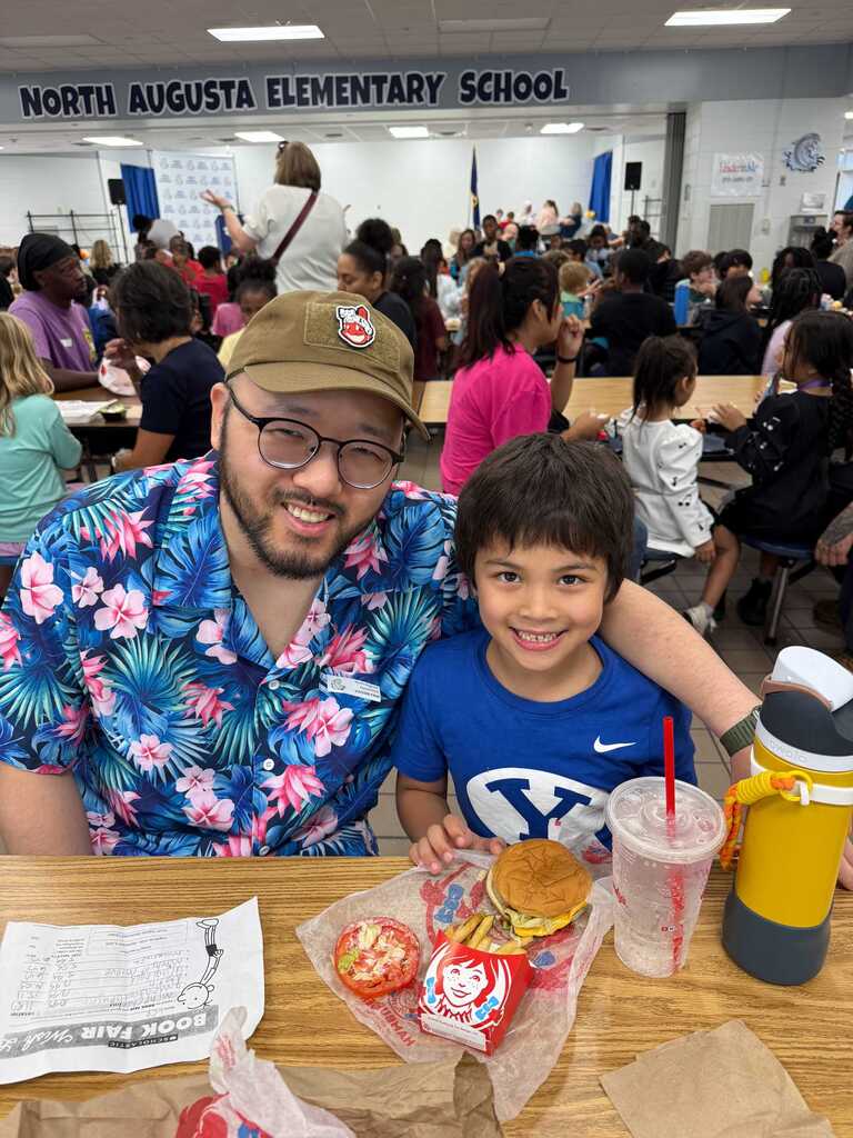 A man and boy sit at a table, smiling. Behind them, a room with many people seated.