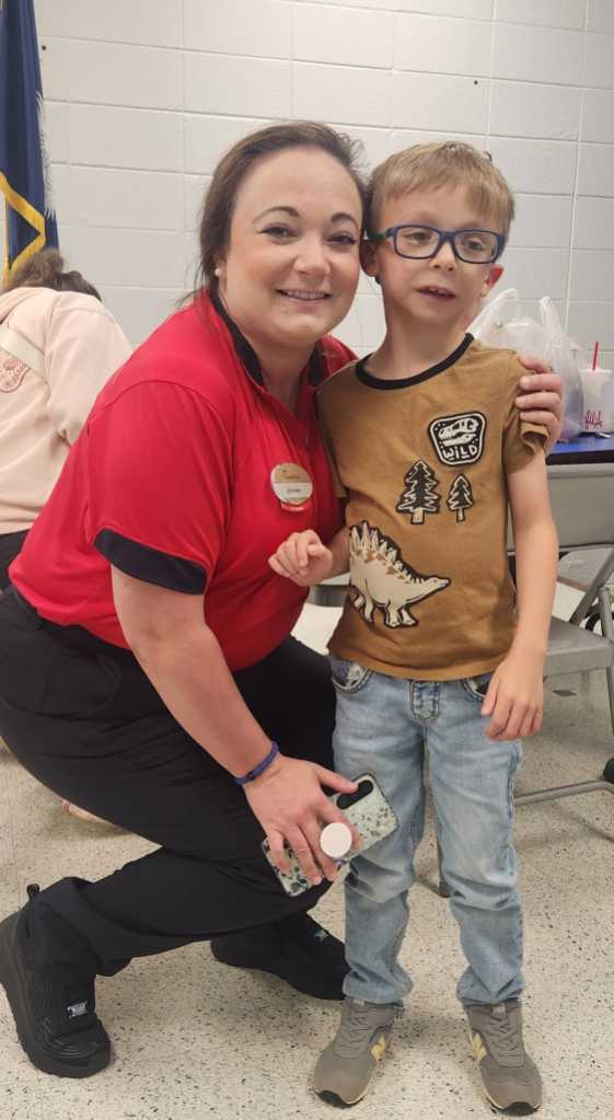 Woman wearing red shirt and black pants kneeling with a boy in glasses and jeans in a room.