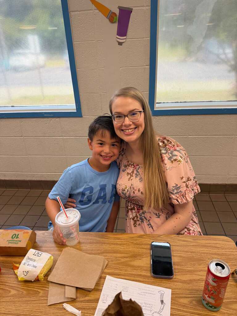 Woman and young boy sit at a table, smiling. A cellphone and paper cup are on the table.