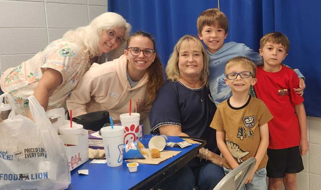 A family gathers around a table with food, drinks, and cups, smiling for a group photo.