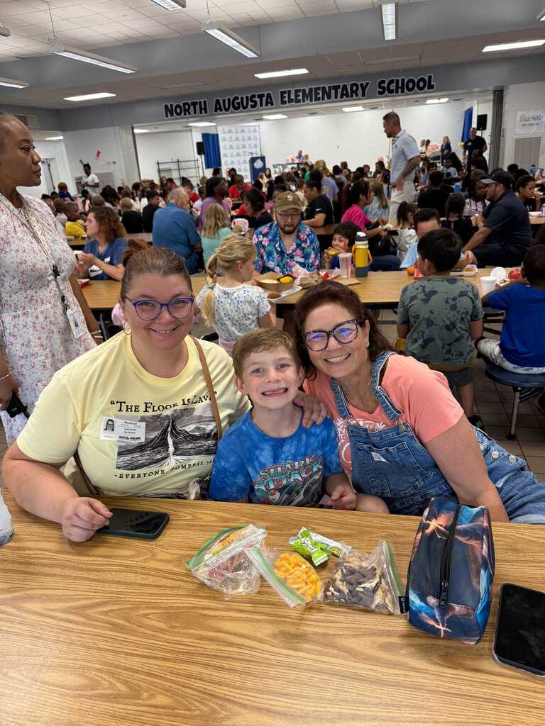 Three people at a table, two wearing glasses, with a child in the middle smiling, inside a school cafeteria.