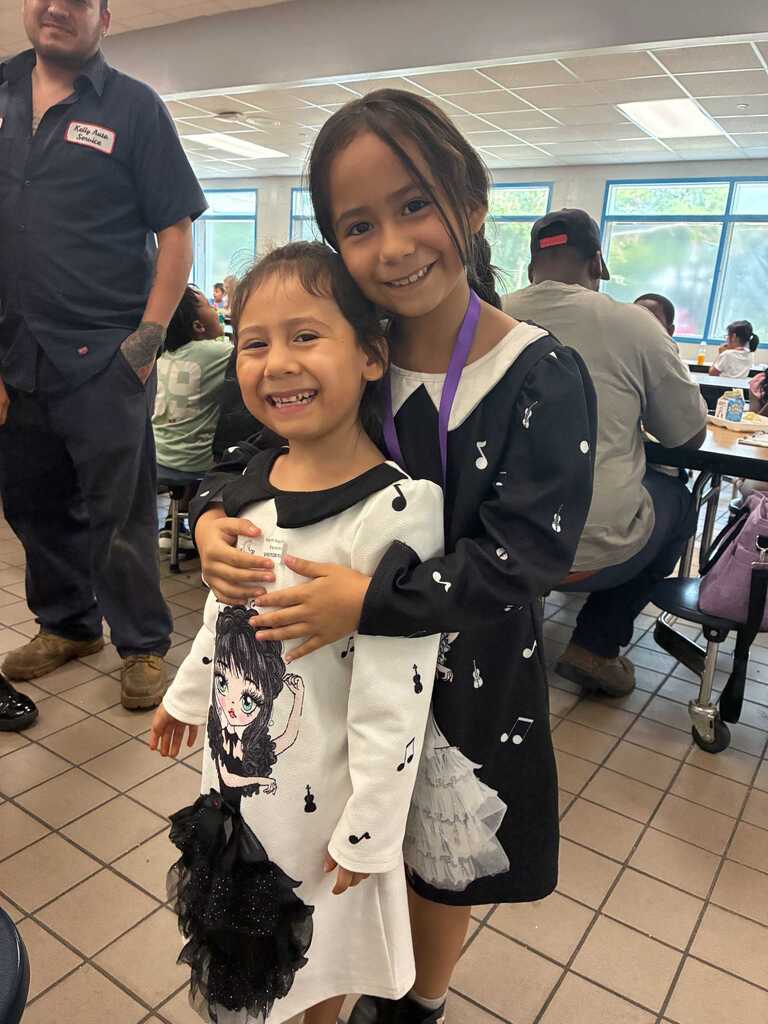 Two children smile, one in a white dress, the other in black. They stand close together. Behind them, a person sits at a table.