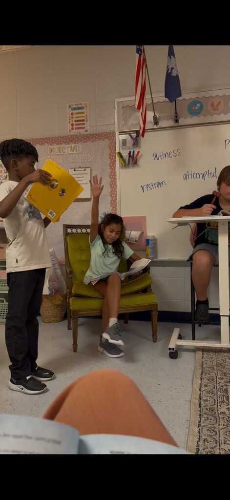 Classroom scene: children sitting on a green chair and desk, holding books, with a whiteboard in the background.
