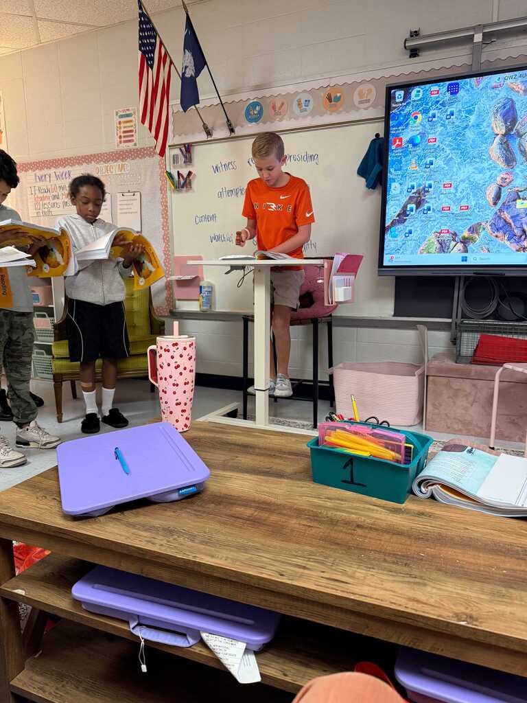 Children in a classroom, one standing at a raised desk, with a whiteboard, flags, and a projector screen behind them.