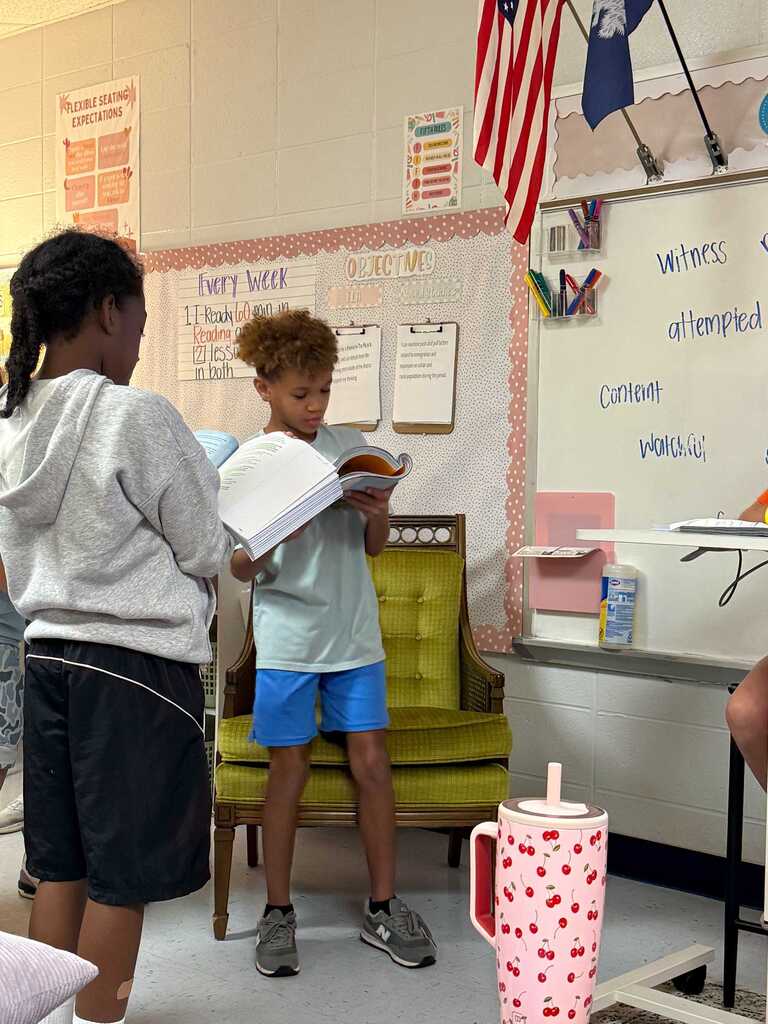 Two children, one reading aloud from a book, stand in a classroom with a whiteboard and American flag in the background.