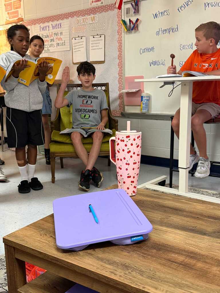 Children in a classroom setting with some seated at a table, others standing. One child holds a book and gestures.