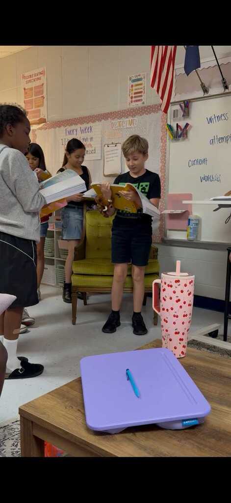 Children stand in a classroom, reading books together. A boy wears a black t-shirt. A girl wears a gray shirt. A table has a purple tray with a pen on it.