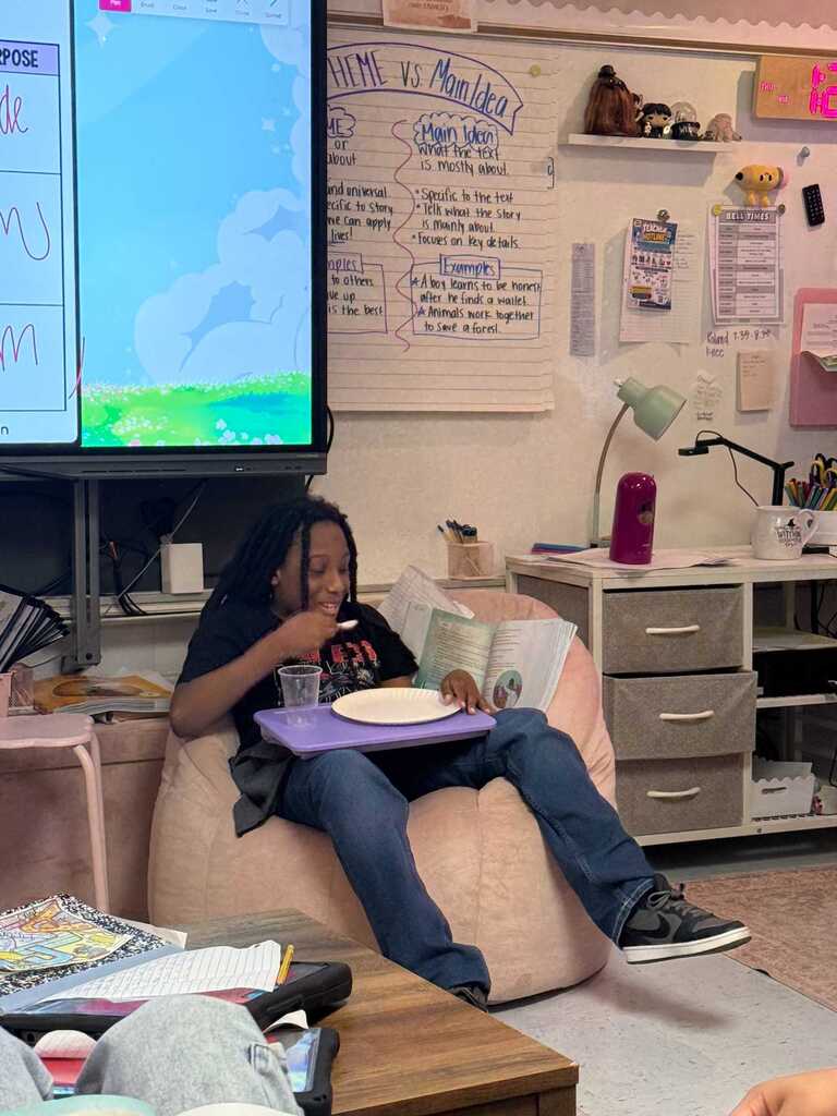 Person in a bean bag chair eating food with a book. A monitor displays a website on the wall.