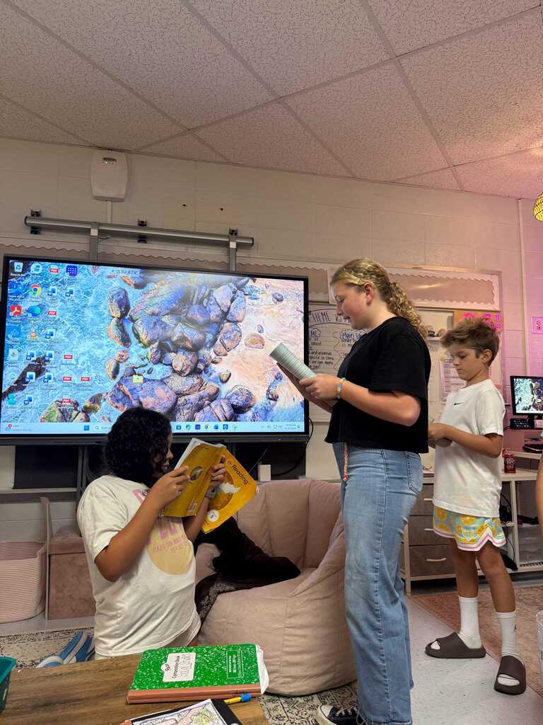 Three kids, one reading a book, one reading from a tablet, and one reading aloud, in a room with a big screen.