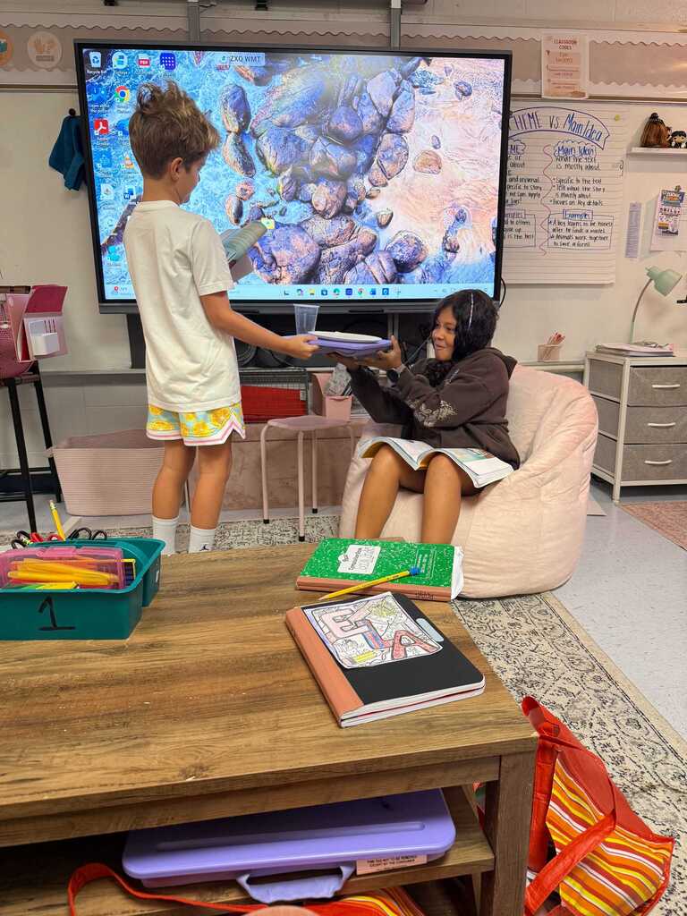 Two children in a room with a large screen displaying an image. One child stands, holding a tablet.