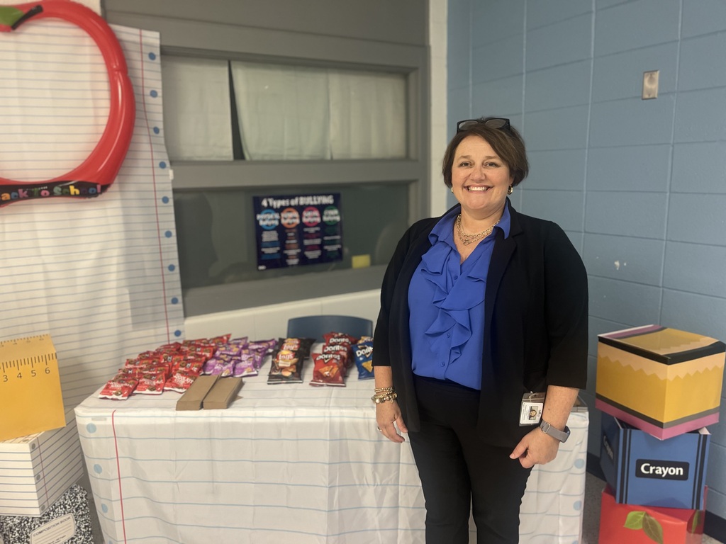 A woman smiles, standing behind a table with snacks. Colorful boxes and a large red circle are in the background.