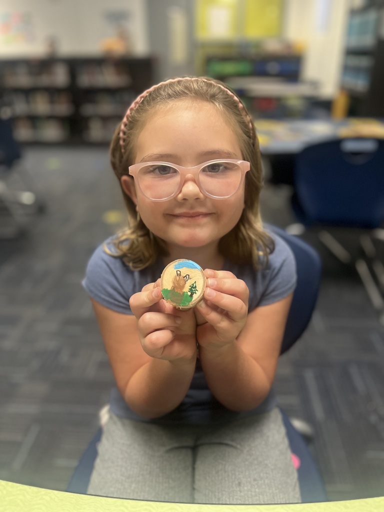 Girl wearing glasses holds a small round object with an animal on it. She is in a classroom with chairs and tables in the background.