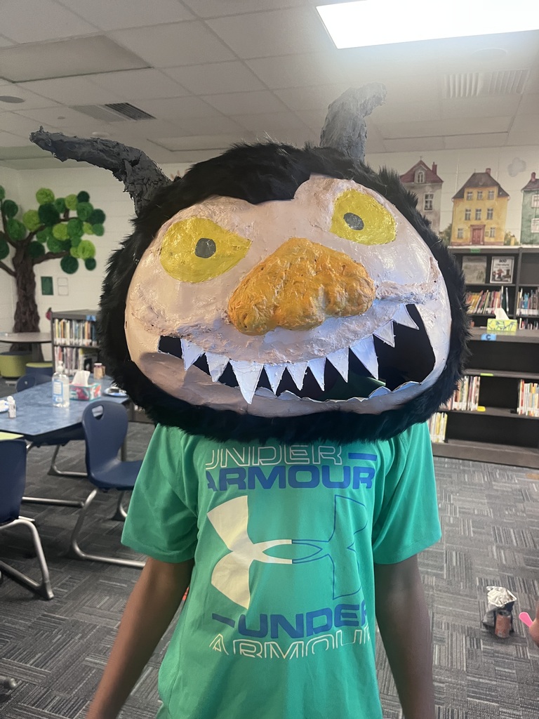 A person wears a large, paper-mache wolf mask with a black background and yellow eyes. They stand in a library with bookshelves and chairs.