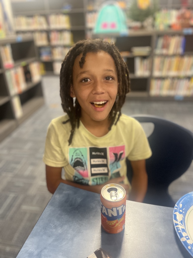 A child with braided hair sitting at a table with a can of Fanta, in a library.