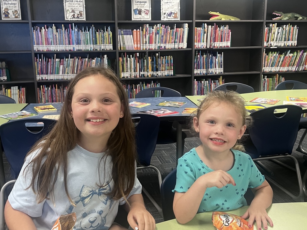 Two children sit at a table in a classroom, smiling and looking at each other. Behind them are bookshelves filled with books.