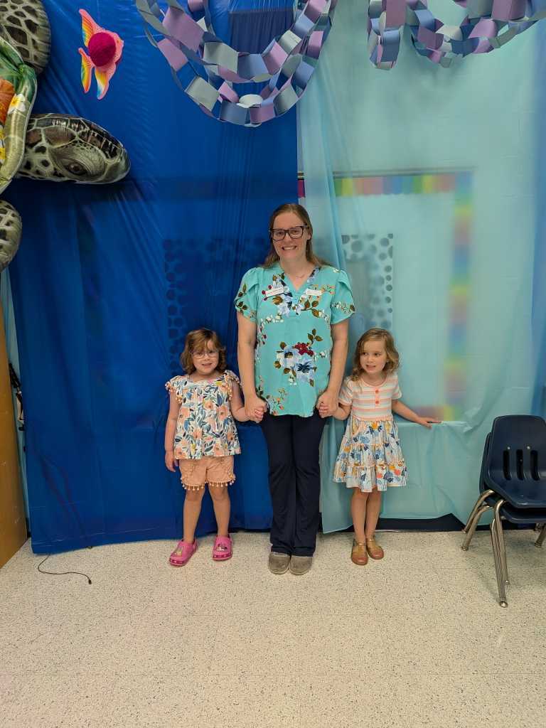 A woman and two girls stand in a room with blue walls, holding hands. The background has a colorful marine life theme.