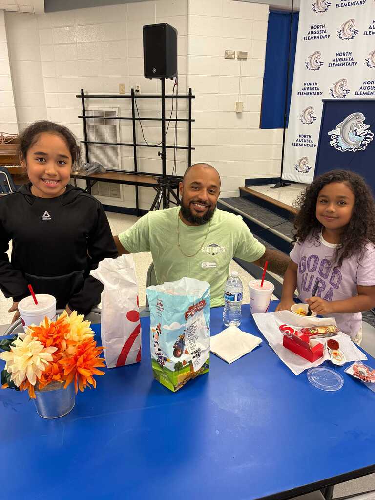 Three people sit at a blue table, smiling. A girl on the left and right hold cups.