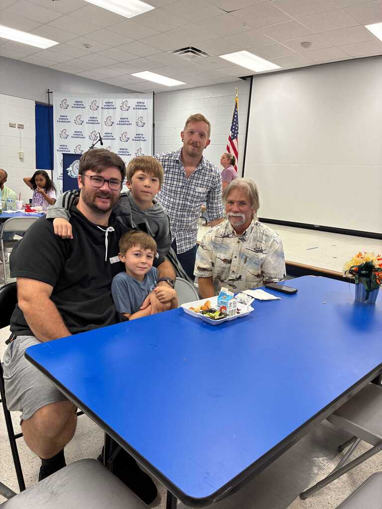 Five people sit at a blue table in a room. A man in a floral shirt is next to a boy in a yellow shirt.