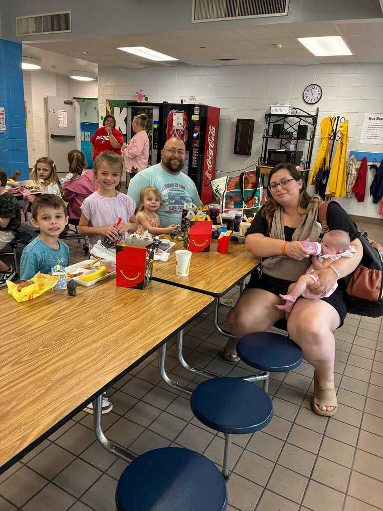 Family gathered at a cafeteria with tables and chairs. A woman holds a baby and sits with children eating.