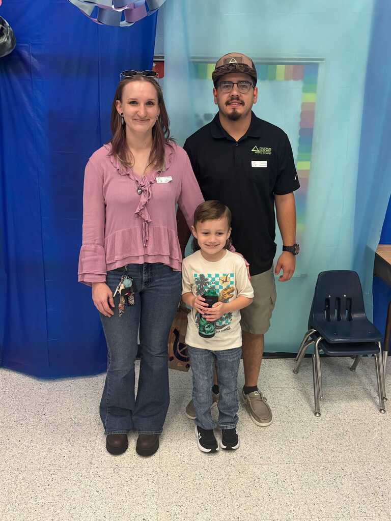 Two adults and a child stand together. The woman wears a pink blouse and jeans, the man a black shirt and shorts. The child holds a toy.