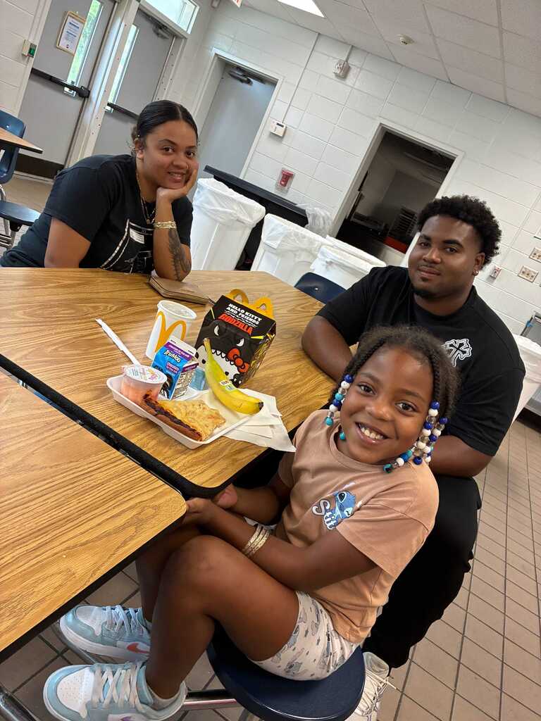 Three people seated around a table with a tray of food and drinks. Two adults and a child.