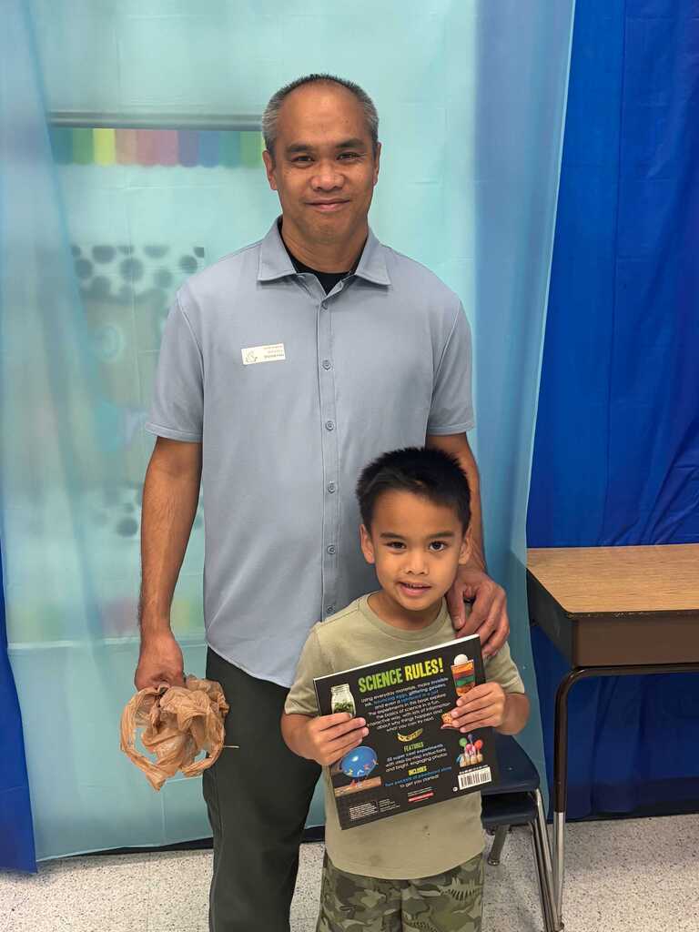 A man stands next to a young boy in a classroom. The boy holds a book and the man holds a wrapped object.