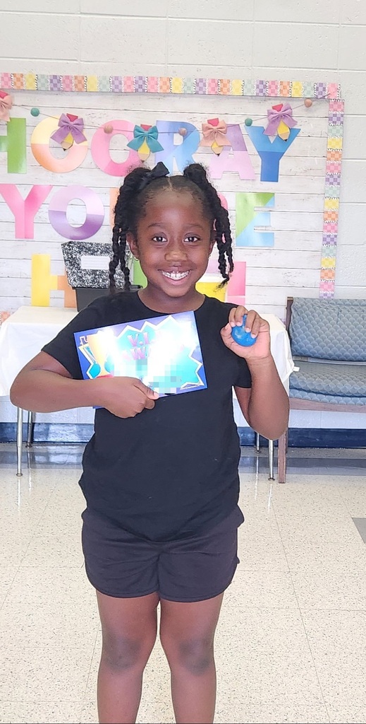A child with braided hair holds a certificate and blue object in a room with a colorful banner.