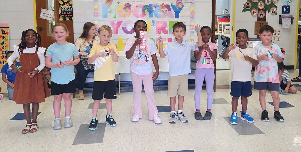 A group of children in a classroom stand in line, each holding a paper object. They wear casual clothing.