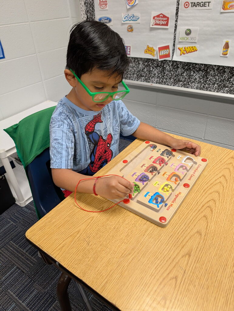 A child wearing glasses sits at a table and works on a wooden puzzle board with colorful characters.