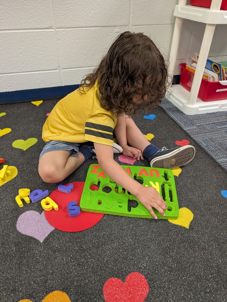 student plays with a green puzzle featuring alphabet pieces, set on a carpeted floor with colorful hearts.