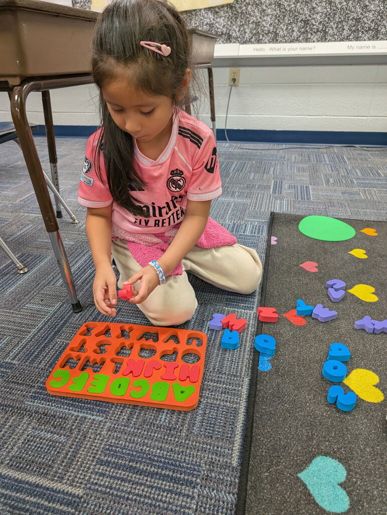 Girl in pink shirt and pants sitting on floor, arranging colorful alphabet tiles on a board.